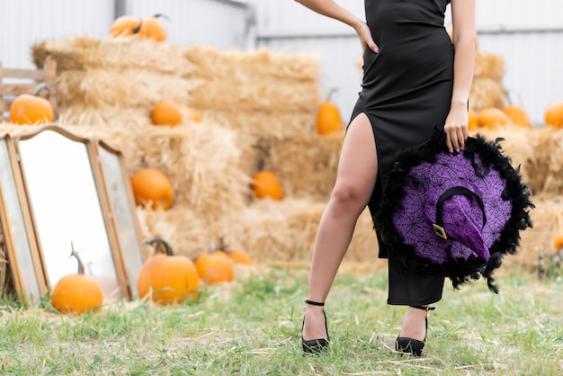 Legs of a beautiful girl in a black dress holds a witch39s hat in his hand posing near an old mirror on a background of hay halloween pumpkin decor