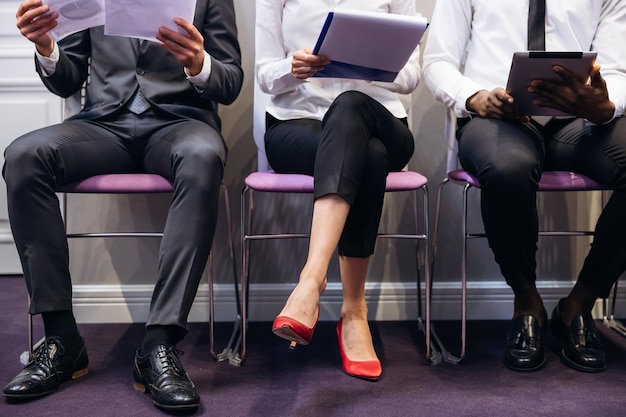 Legs of business people sit on chairs in line for job\
interview