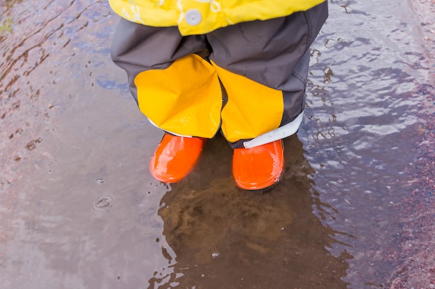 オレンジ色のゴム製のブーツの子供の足は、秋の水たまりにジャンプします。子供の明るいゴム長靴