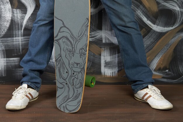 Legs of a fashionable man in jeans and sneakers with a skateboard in his hands closeup Man poses against a painted wall