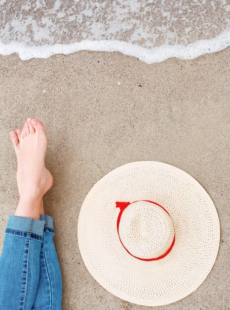 legs and feet of caucasian woman in jeans sitting by the sea on the coast with hat near by on sand