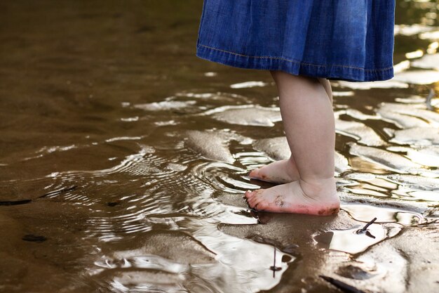 Legs of a little girl in a denim dress standing on the sand of a riverbank in the water in summer. Childhood.