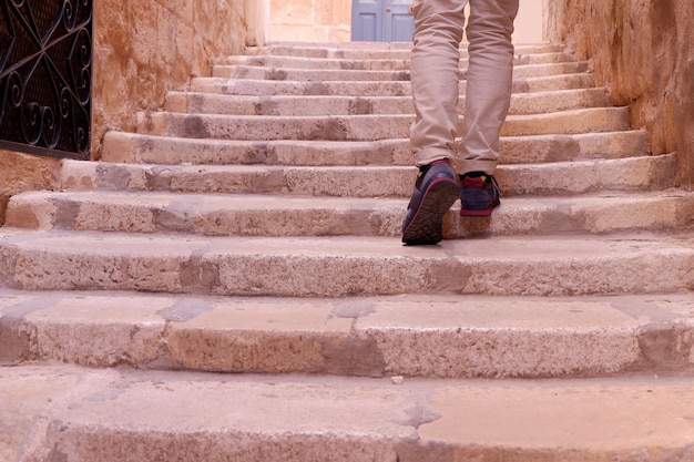 Legs of a man in beige trousers the moment of climbing up the\
stairs to a perpendicular street granite stone of light color birgu\
malta