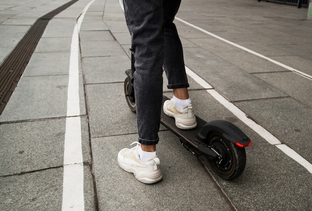 街の通りのぼやけた背景のアスファルトパスで現代の電動スクーターに乗っている男の足