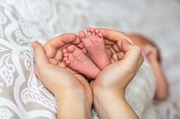 Ноги новорожденного в руках родителей на белом фоне. Маленькие ножки новорожденного в большой руке взрослого. Ноги новорожденного в руках матери на одеяле светлом фоне.