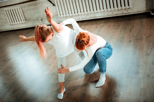 Legs position. Professional modern dance teacher with red hair wearing jeans correcting legs position of her student