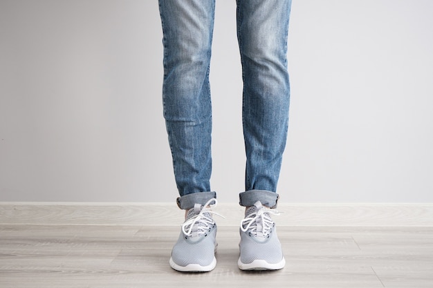 Legs of a young man in jeans and sneakers on gray wall.