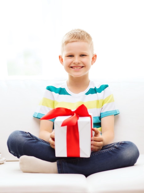 Photo leisure, childhood, holidays and home concept - smiling little holding gift box with red bow and sitting on couch