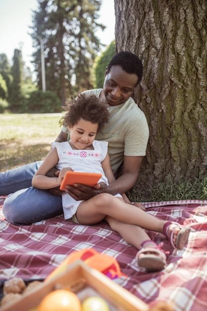 Photo leisure. curly kid spending time with her dad and watching something on a tablet