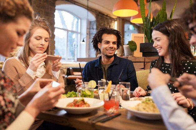 레저, 음식, 음료, 사람 및 휴일 개념 - 스마트폰을 가진 행복한 친구들이 레스토랑에서 먹고 마시고 있습니다.