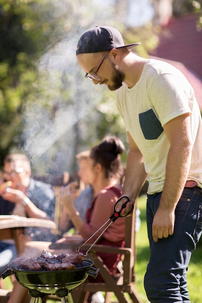 写真 レジャー、食べ物、人、休日のコンセプト – 夏の屋外パーティーで友達のためにバーベキューグリルで肉を料理する男性