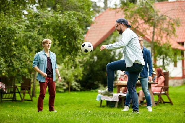 концепция отдыха, праздников, людей и спорта - счастливые друзья играют в футбол на летней вечеринке в саду