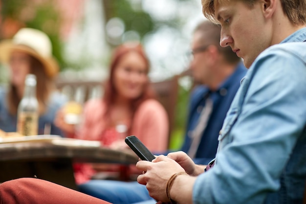 leisure, holidays, people and technology concept - young man texting on smartphone and friends having dinner at summer garden party