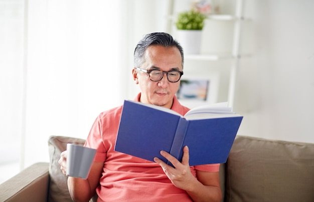 leisure, literature and people concept - man sitting on sofa with mug and reading book at home