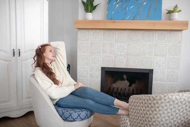 Photo leisure. pretty woman touching long ginger hair sitting in chair relaxing at home near fireplace