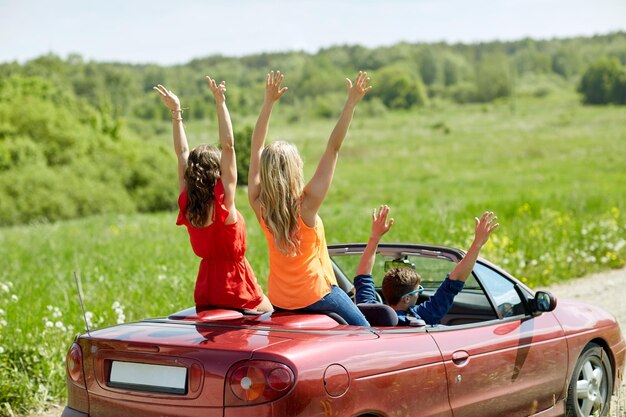 Concetto di tempo libero, viaggio su strada, viaggio e persone - amici felici che guidano in auto cabriolet in campagna e agitano le mani