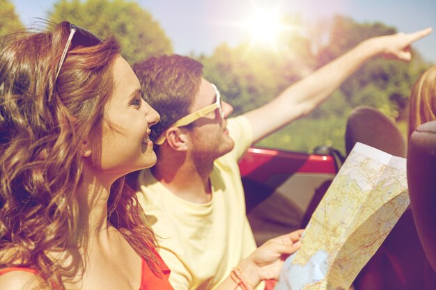 leisure, road trip, travel and people concept - happy friends with map driving in cabriolet car at country and pointing finger to something ahead