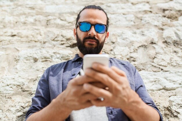 leisure, technology, communication and people concept - man in sunglasses texting message on smartphone at stone wall