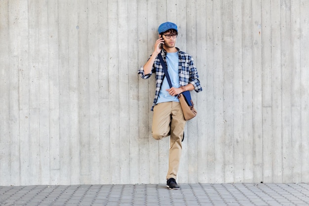 leisure, technology, communication and people concept - smiling hipster man with shoulder bag calling on smartphone at street wall