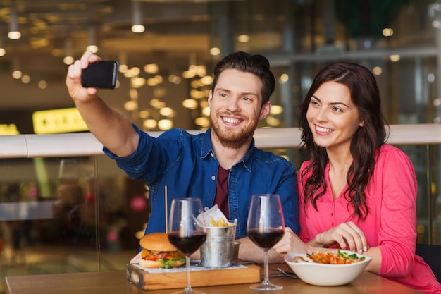 レジャー、テクノロジー、デート、人、祝日のコンセプト – 幸せな夫婦がレストランで夕食をとり、スマートフォンで自撮りをする