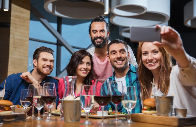 leisure,technology, friendship, people and holidays concept - happy friends having dinner and taking selfie by smartphone at restaurant