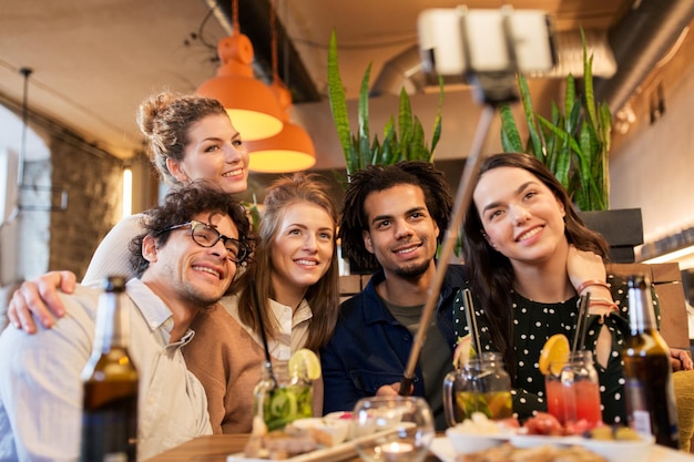 leisure, technology, friendship, people and holidays concept - happy friends with food and drinks taking picture by smartphone selfie stick at bar or cafe
