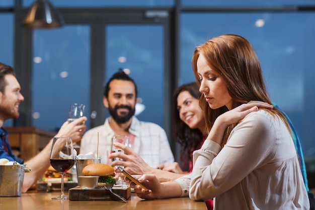 leisure, technology, internet addiction, lifestyle and people concept - woman with smartphone and friends at restaurant