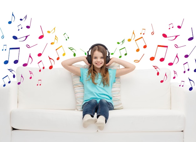 Photo leisure, technology, music and childhood concept - smiling little girl in headphones listening to music sitting on sofa over musical notes