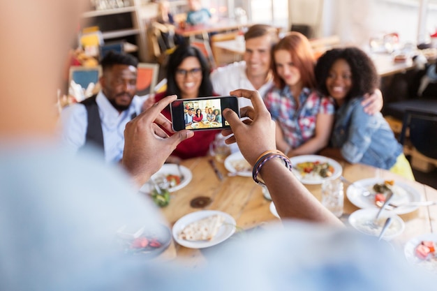 レストランのテーブルでスマートフォンで食べたり写真を撮ったりする 幸せな国際的な友達のグループ