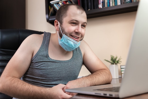 Leisure using laptop. A young bearded guy with face mask in home clothes uses laptop 