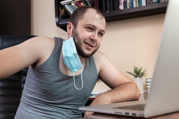 Leisure using laptop. A young bearded guy with face mask in home clothes uses laptop 