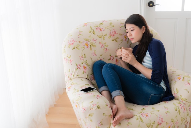 快適なソファの椅子に座ってカップを飲みながら、良いメッセージを期待して携帯電話を見ているのんびりした美人女性。