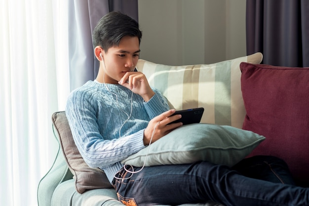 Leisurely day of young Asian man wearing headphones watching their phone sleep at sofa at home.