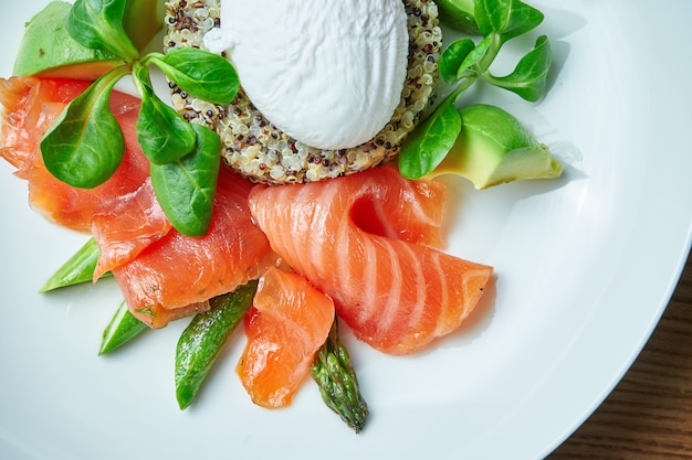 Lekker en gezond continentaal ontbijt - quinoa met benedict eieren, spinazie, avocado, gezouten zalm en asperges in witte kom. Houten tafel