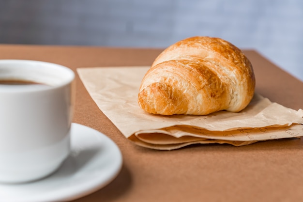 Lekker ontbijt. De Franse croissant diende op ambachtdocument en kop van zwarte koffie of espresso op bruine achtergrond. banier