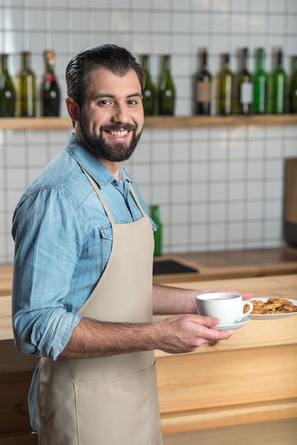 Lekker ontbijt. Professionele vrolijke jonge ober die er vriendelijk uitziet terwijl hij in de buurt van de bar staat en een kop warme koffie en een bord met lekkere koekjes in zijn handen houdt