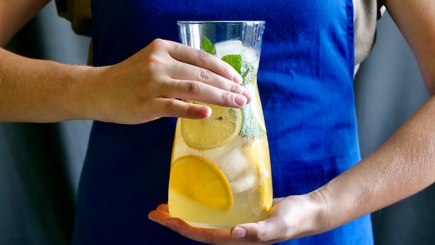 Лимонный лимонад с мятным лимоном и льдом в графине Женщина в синем фартуке держит прохладительный освежающий фруктовый летний напиток