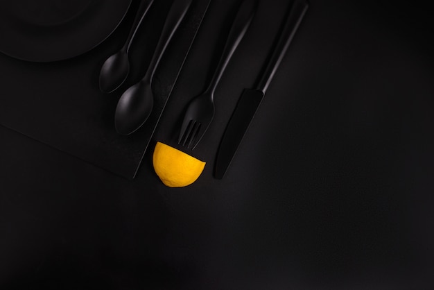 Lemon slices on a black background with black cutlery, a black glass and a black plate, top view