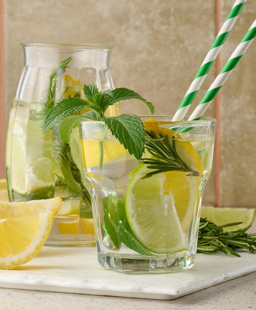 Lemonade in a transparent glass with lemon lime rosemary sprigs and mint leaves on a white background