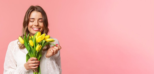 Lente geluk en viering concept close-up portret van mooie romantische blond meisje snuiven geur van mooie gele tulpen ogen sluiten en glimlachend gelukkig permanent roze achtergrond