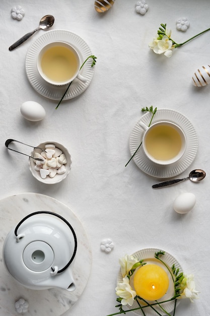 Lente middagthee Lente fresia bloemen theepot en kopje thee op tafel Plat leggen van wit textiel tafelkleed Paaseieren witte keramische kopjes en suikerharten