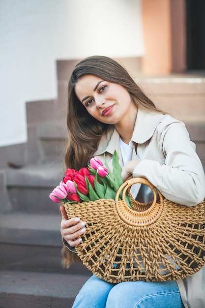 Lente portret van een jonge mooie gelukkige vrouw 28 jaar oud met lang goed verzorgd haar houdt een rieten tas in haar handen met een boeket tulpen op een stadsstraat Stijlvol model in een trenchcoat