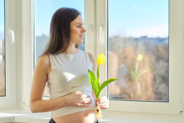 Lente, schoonheid, gezondheid, portret van jonge zwangere vrouw met gele tulp thuis dichtbij venster. Mooie vrouw met een gezonde huid en haren, natuurlijke make-up, kopie ruimte