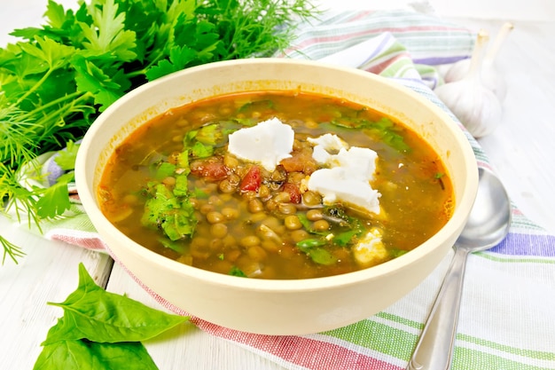 Lentil soup with spinach tomatoes and feta cheese in a yellow bowl spoon on a kitchen towel parsley and spinach on the background light wooden boards