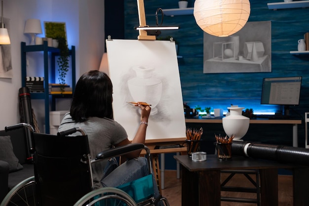 Leraar beeldende kunst die leeft met een handicap in de benen legt de laatste details op kunstwerken op canvas in een tekenworkshop. afro-amerikaanse vrouw met talent voor schetsen, rondhangen in de kunststudio thuis.