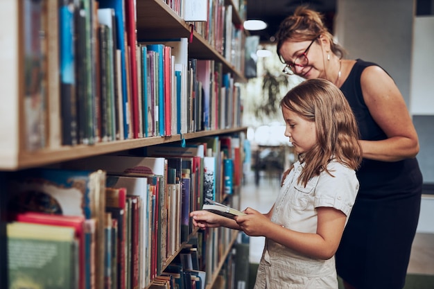 Foto leraar helpt bij het kiezen van haar boek schoolmeisje in de schoolbibliotheek meisje selecteert boeken om te lezen