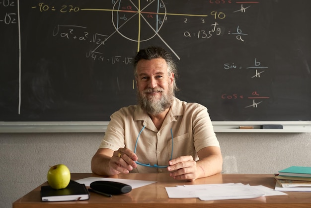 Leraar met vriendelijk gezicht kijkend naar de camera zittend aan zijn bureau in de klas met de zwarte