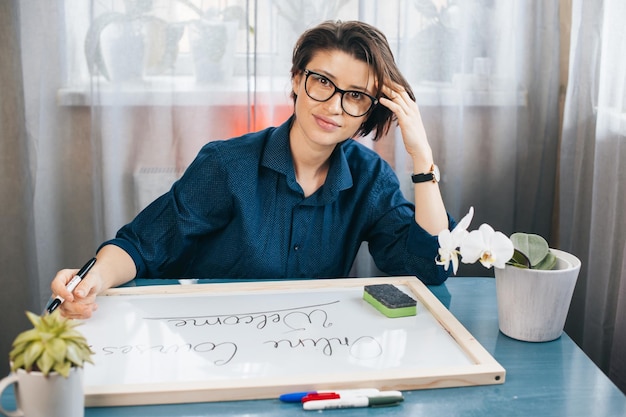 Leraar poseren zittend aan een bureau kijkend naar de camera die zinnen schrijft op een wit bord met blauwe markering