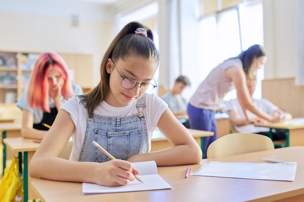 Les in de klas van tienerkinderen vooraan meisje 13 14 jaar oud zittend aan bureau schrijven in notitieblok Onderwijs school college schoolkinderen concept