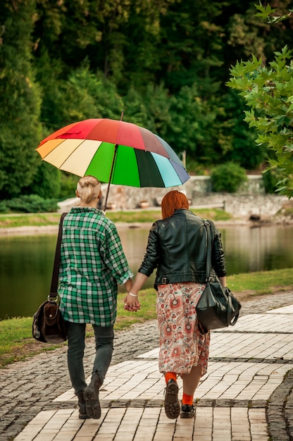 手を繋いでいるレズビアンのカップルは、虹の傘で雨の中で秋の公園を散歩します。